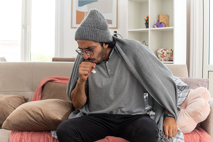 Man wearing a gray beanie and glasses coughing into his hand, wrapped in a blanket on a living room couch feeling unwell.