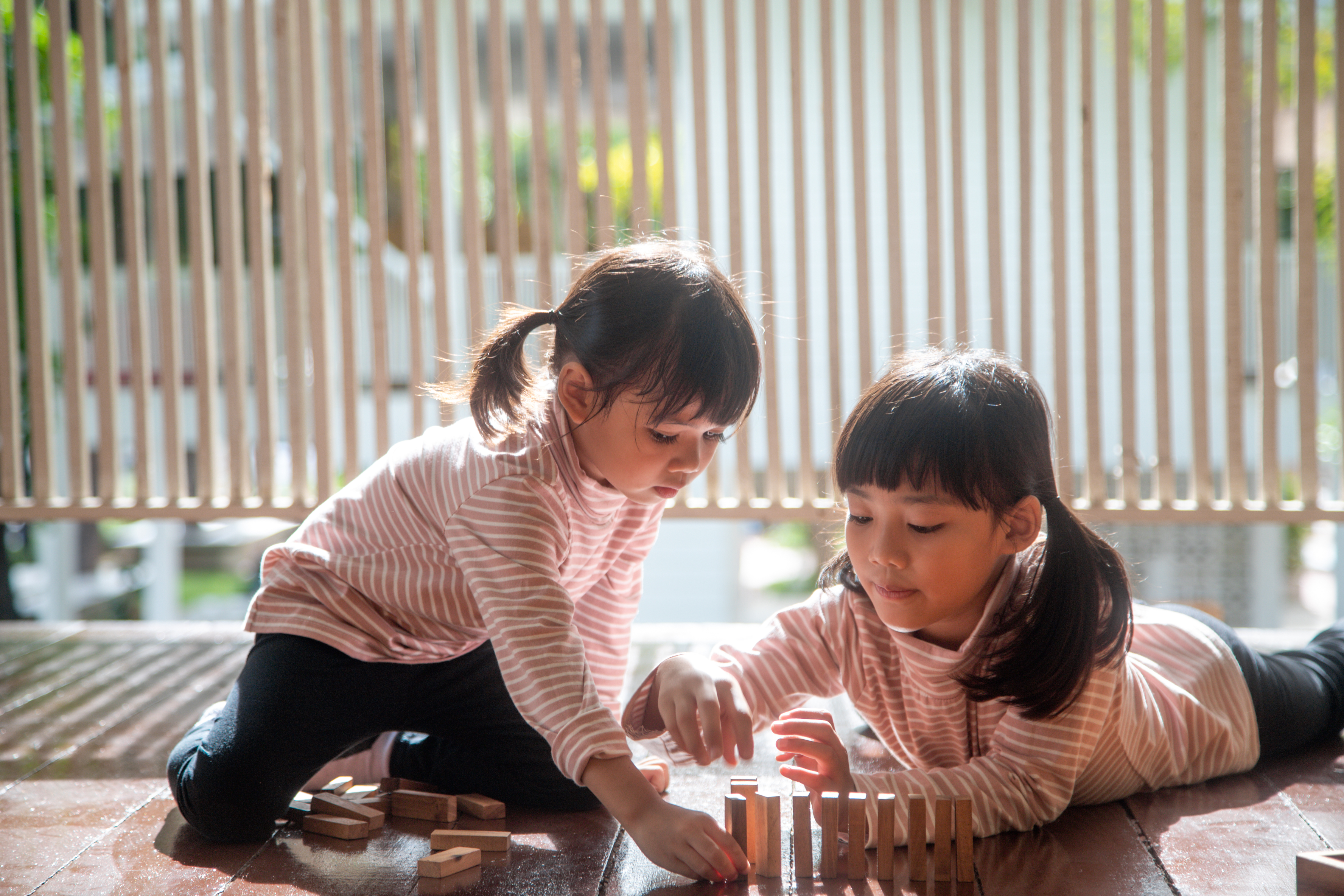 Two young best friend girls in pink striped shirts play with wooden blocks on a sunny floor.