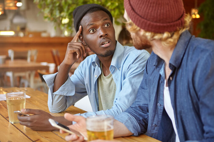 Two men at a cafe, one looking surprised while listening to a friend, discussing stories about being cheated on.