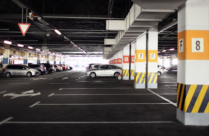 A bright, clean underground parking lot with parked cars, marked arrows on the asphalt, and numbered yellow and black striped pillars, like messy anonymous confessions.