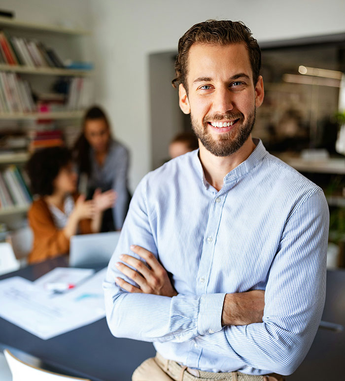 Smiling young professional standing confidently in office with team collaborating in the background demonstrating creative ways to get a job.