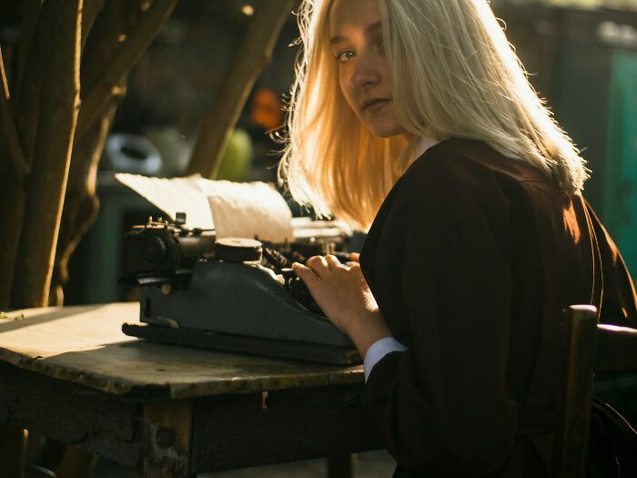 Young woman with blonde hair sitting at a wooden table using an old typewriter, illustrating karma stories concept.