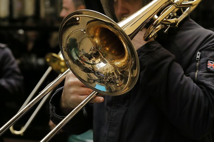 Person playing a trombone outdoors with reflections visible on the brass instrument’s bell.