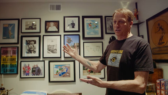 A man in a Birdhouse shirt gestures in a room with a gallery wall. He shares celebrity relatives stories.