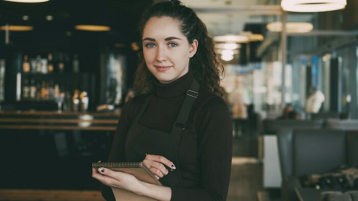 Young woman with curly hair and apron holding notepad in cozy restaurant, representing jobs AI can’t replace.