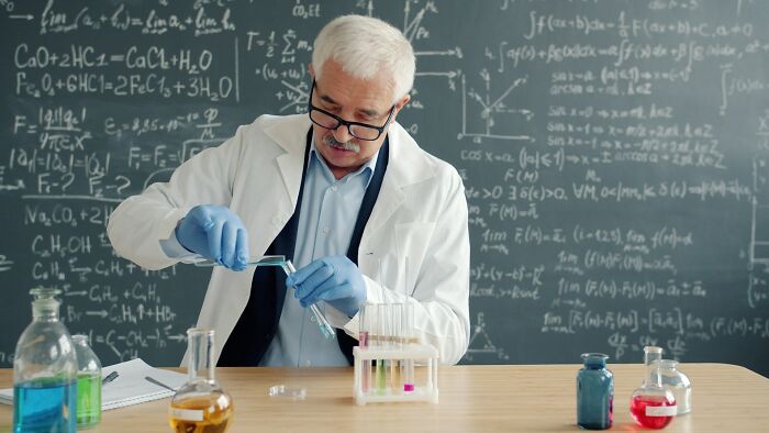 Older male scientist wearing glasses and gloves, conducting chemistry experiments in a lab with formulas on a blackboard.