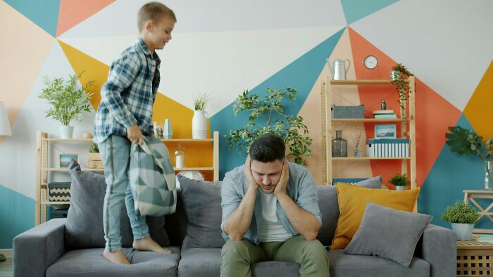 A boy jumps on a couch while his father covers his ears, looking stressed. Highlights family secrets.
