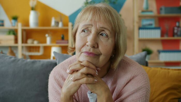 A senior woman in a pink sweater, thoughtfully looking away, her hands clasped. MIL stories keyword.