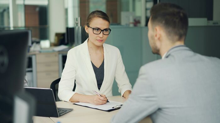 Woman in a white blazer and glasses interviewing a man in a gray suit, highlighting encounters with former bullies.