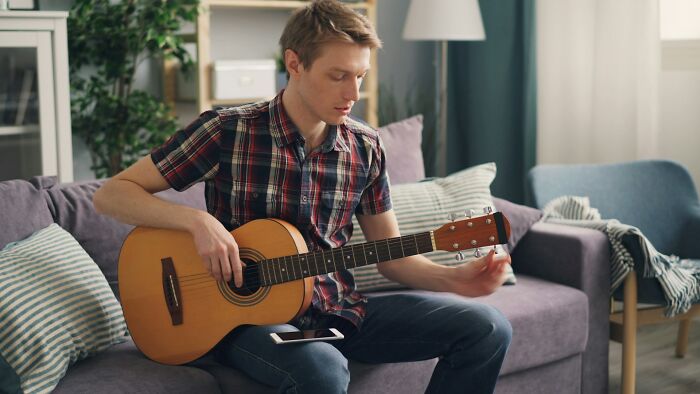 Young man playing acoustic guitar on a couch, demonstrating intelligence in a casual home setting with natural light.