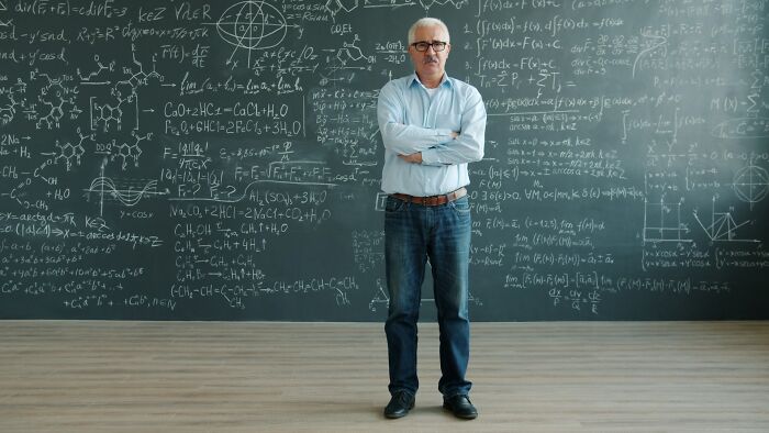 Older man standing confidently in front of a chalkboard filled with complex mathematical formulas, illustrating karma concepts.