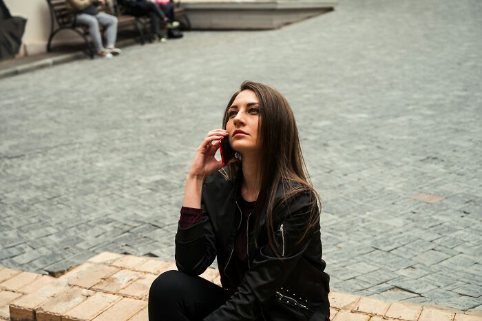 A young woman with long brown hair, sitting on stone steps, talking on a red phone. She looks thoughtful, perhaps about to rage quit her job.