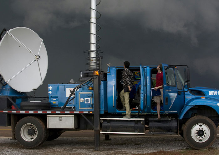 Scientists standing by a blue research truck with satellite equipment, studying weather phenomena and unproven scientific theories.
