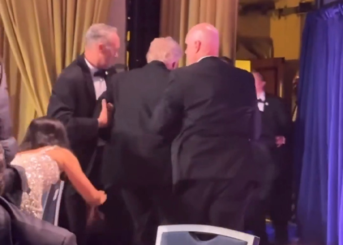 California teacher escorted from the White House Correspondents Dinner. A man in a tuxedo pulls another man, in front of a woman in a sparkly dress.