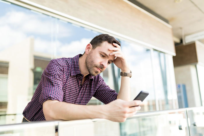 Man moves out to save marriage, looking stressed while checking his phone on a balcony with glass railing background.