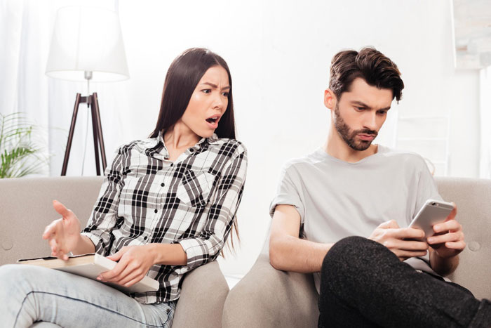 Couple having intense argument on couch, woman shocked, man focused on phone, illustrating marriage and bedroom twist tension.