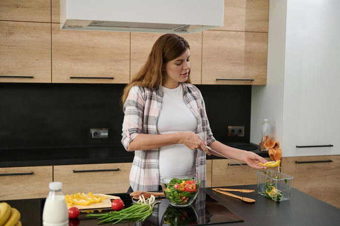 Pregnant woman preparing salad in kitchen, reflecting on wife&rsquo;s career glow-up and marriage trouble worries.