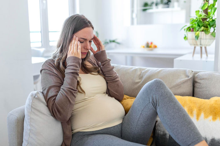 Pregnant woman sitting on couch, stressed and worried about marriage trouble amid wife&rsquo;s career glow-up.