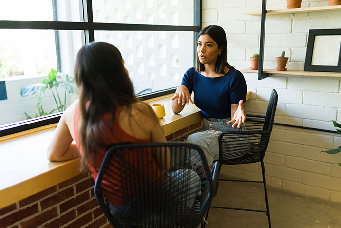 Two women having a serious conversation by a window in a cafe, capturing tension and emotional conflict.