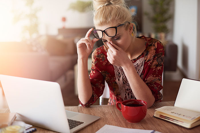 Blonde woman looking stressed, wearing glasses and floral shirt, laptop and coffee nearby. She finds husband secret wife.