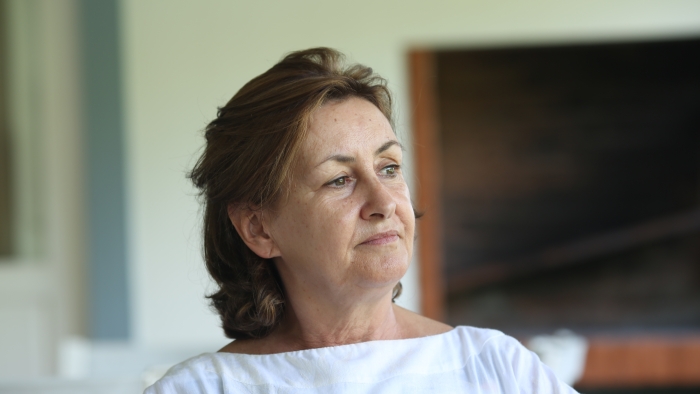 Uma mulher pensativa, de cabelos castanhos, de camisa branca, desviando o olhar. Concentra-se no segredo e na infidelidade da MIL.