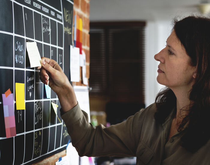 A woman organizes a blackboard calendar with sticky notes, representing "cheat codes" for surviving adult life.