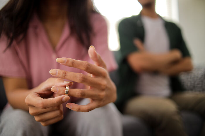 A person's hands removing a wedding ring, while another person sits blurred in the background, depicting being cheated on.