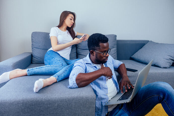 A woman on a sofa looks at her phone while a man works on a laptop, unaware he's being cheated on.