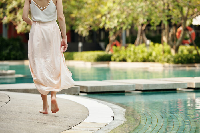 A woman in a flowing skirt walking barefoot along a pool's edge. She is finding out she was cheated on.