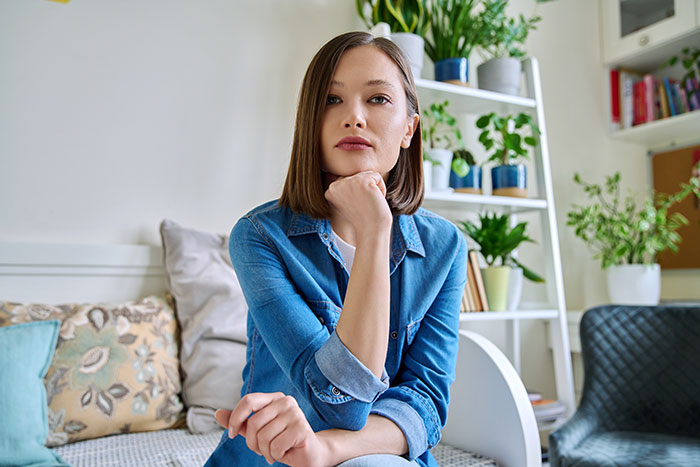 A thoughtful woman in a denim shirt, resting her chin on her hand, surrounded by houseplants. Boyfriends and husbands can be so mean.