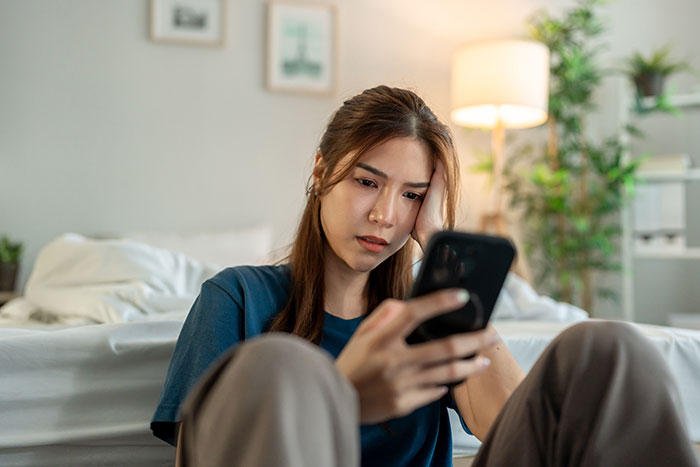 A young woman with a frustrated expression, head in hand, looking at her phone. She is venting online about boyfriends.
