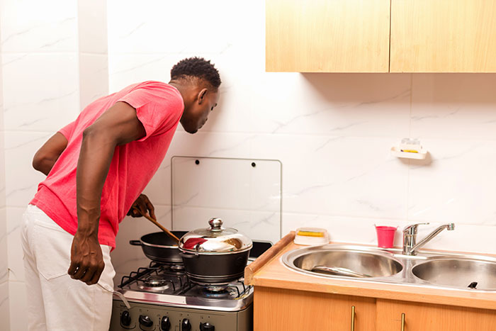 A man in a red shirt and white pants cooks on a stove, stirring a pot. Could he be a mean boyfriend or husband?