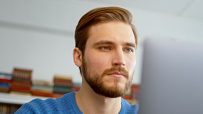 A focused man with a beard and blue eyes, wearing a blue sweater, looking at a screen. He might be one of the boyfriends.