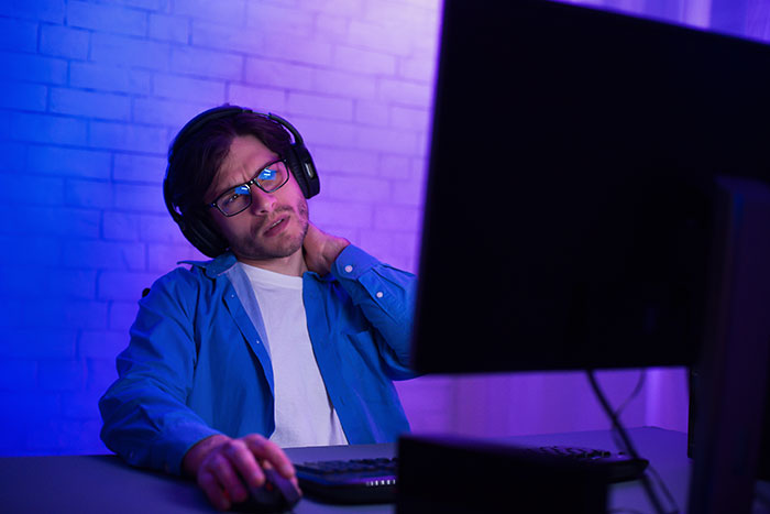 A male gamer with headphones and glasses rubbing his neck in pain while using a computer. Boyfriends and husbands often cause frustration.