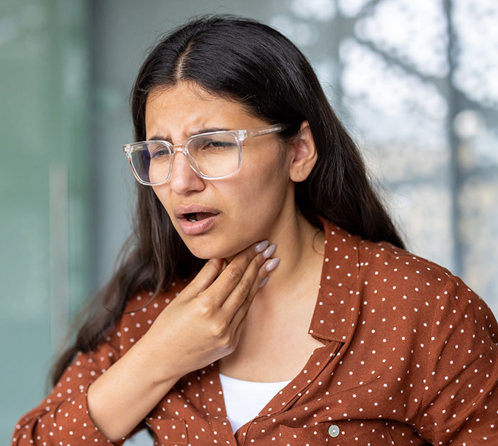 A woman with glasses and a brown polka-dot shirt holding her throat, looking distressed by mean boyfriends and husbands.