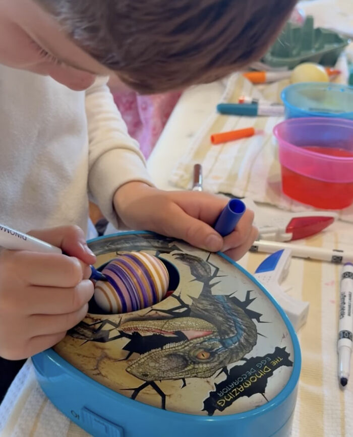 Child decorating Easter egg using a blue marker with dinosaur egg color kit at a crowded craft table.
