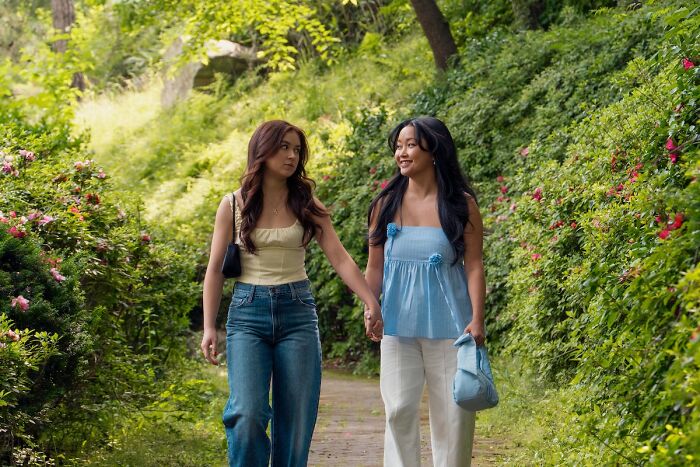 Two young women holding hands and walking on a path surrounded by greenery in XO Kitty season 3 scene.
