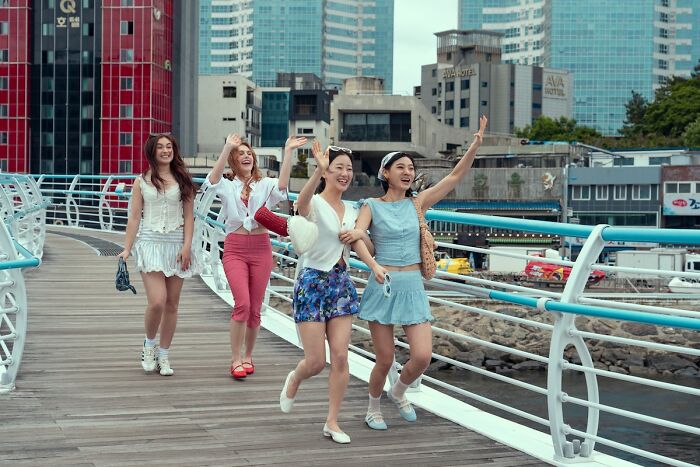 Four young women happily walking and waving on a city boardwalk, capturing XO Kitty tension in messy season 3 atmosphere.