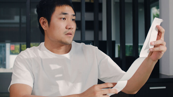 A man in a white t-shirt looks shocked while reading a long paper, perhaps finding out he was being cheated on.