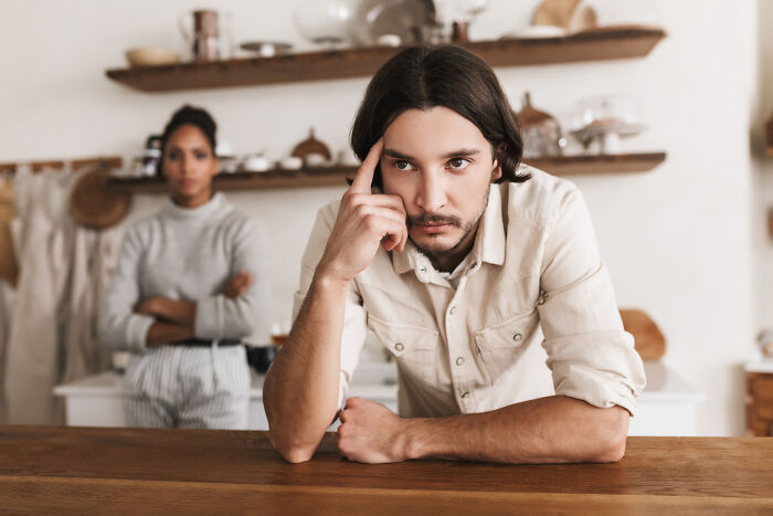A pensive man with long hair and a goatee, leaning on a counter, thinking about being cheated on. A woman stands in the background.