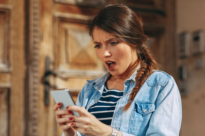 A surprised woman with an open mouth, wearing a denim shirt, looking at her phone. She is accidentally finding out she was being cheated on.