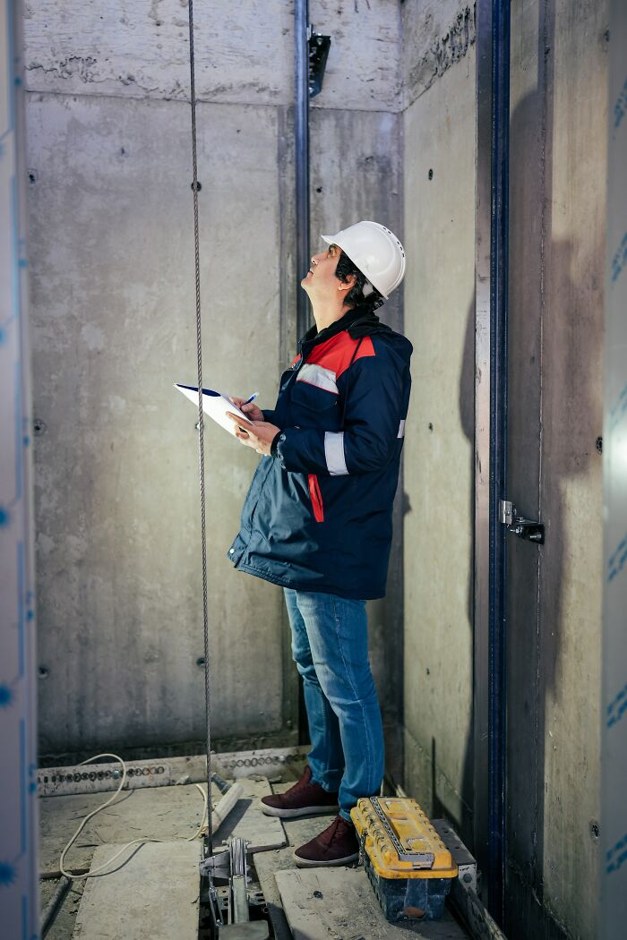 Elevator technician inspecting elevator pit, wearing a hard hat and jacket, holding a clipboard during maintenance check.