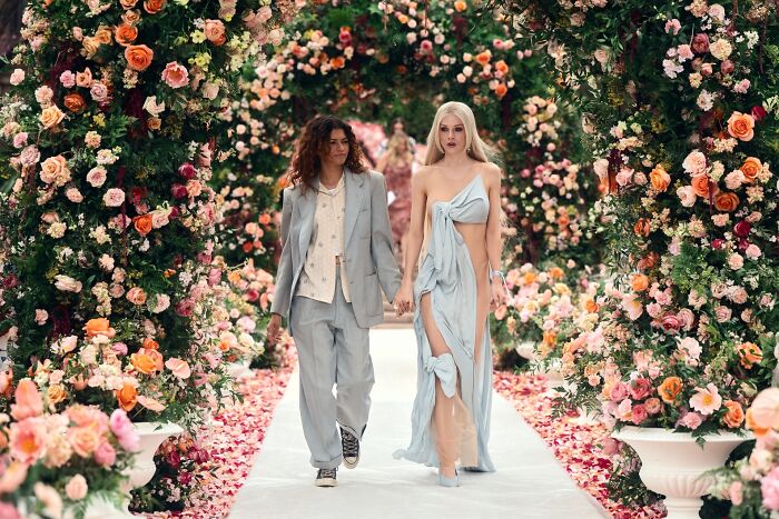Two actresses from Euphoria, Sydney Sweeney and Zendaya, walk hand-in-hand down an aisle adorned with flowers.