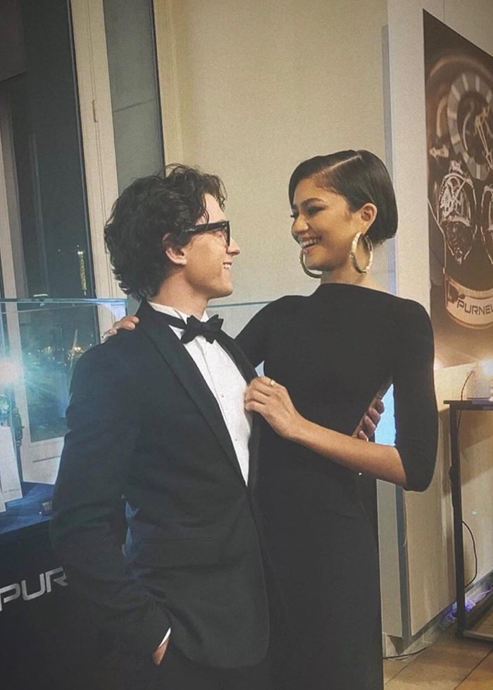 Zendaya in elegant black sheer dress with hoop earrings smiling at man in tuxedo during movie promo event.