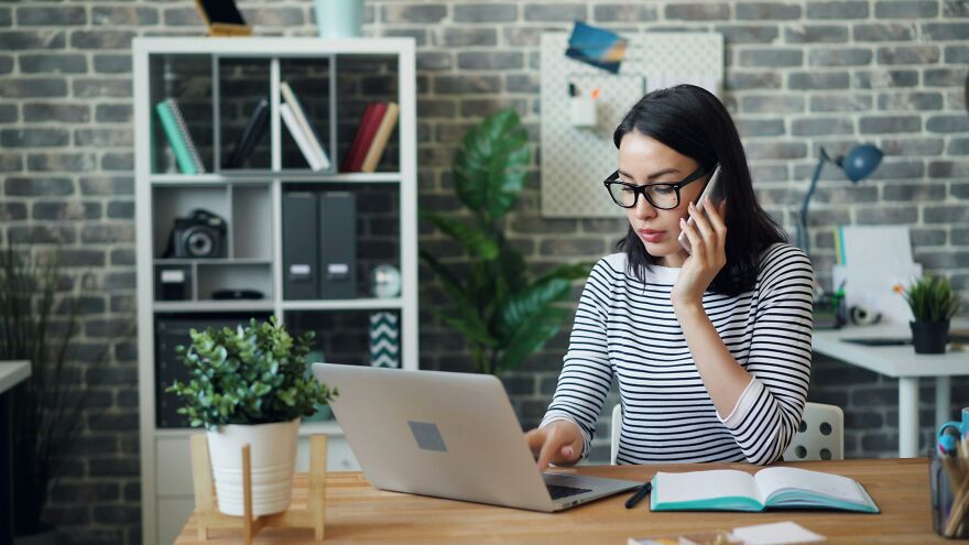 A woman in glasses and a striped shirt talks on the phone while typing on a laptop. She faces the consequences of liars getting caught.