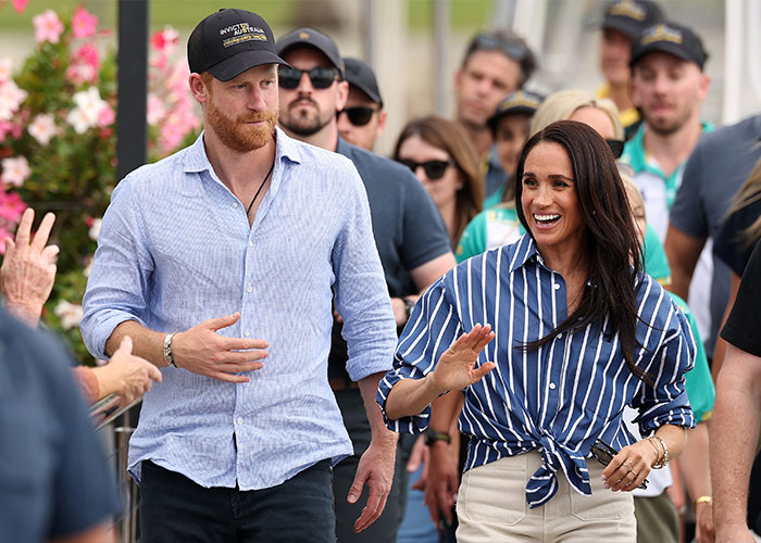 Meghan Markle and Prince Harry amidst a crowd, Markle smiling and waving. Scrutiny on Meghan Markle's necklace. Meghan Markle and Prince Harry amidst a crowd, Markle smiling and waving. Scrutiny on Meghan Markle's necklace.