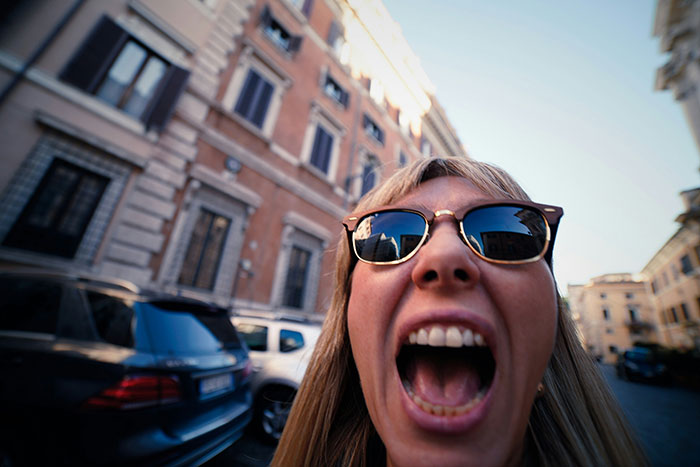 A woman with sunglasses, mouth wide open, screaming in front of buildings. Her honeymoon behavior is questionable.