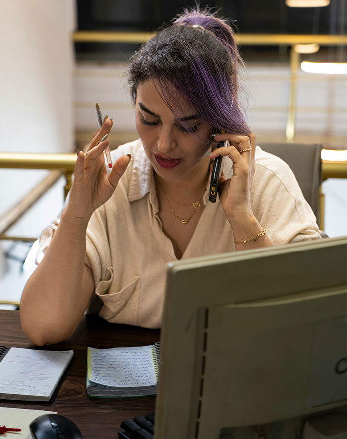 A woman with purple-streaked hair, a school administrator, talking on the phone at her desk, holding a pen, expressing a cathartic moment. A woman with purple-streaked hair, a school administrator, talking on the phone at her desk, holding a pen, expressing a cathartic moment.