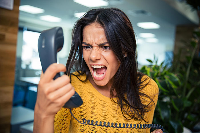 A woman with dark hair in a yellow top screams into a phone, expressing cathartic anger, possibly a school administrator dealing with an entitled mom. A woman with dark hair in a yellow top screams into a phone, expressing cathartic anger, possibly a school administrator dealing with an entitled mom.