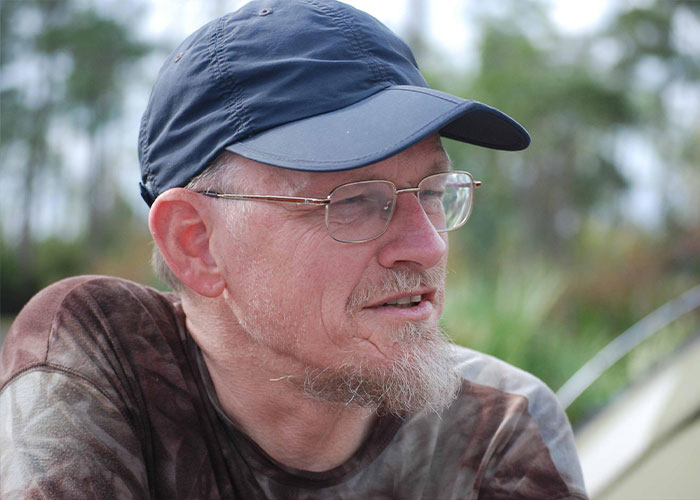 Man in a blue cap and glasses, with a goatee, possibly Patient Zero, in an outdoor setting.